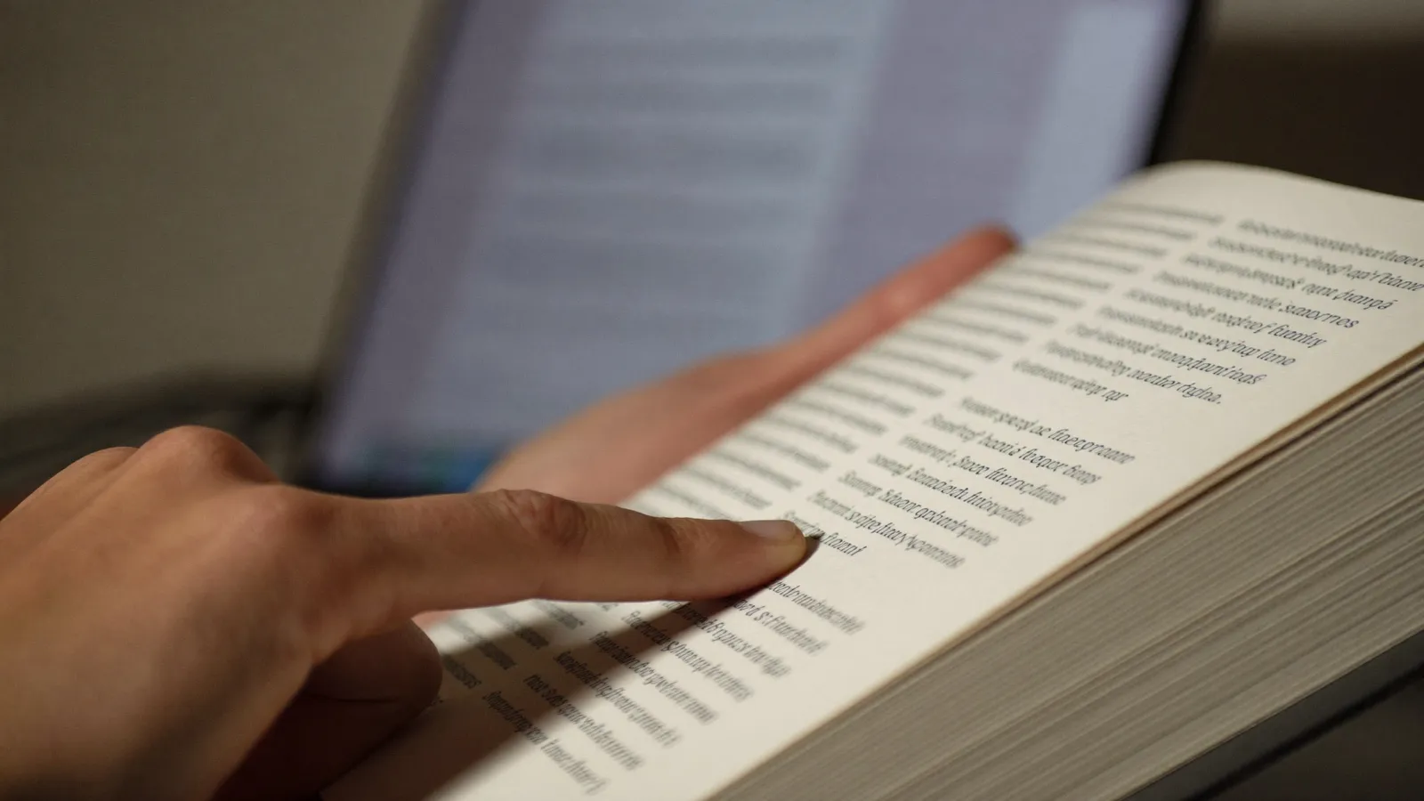 A finger resting on a line of text in an open book, with the glow of a laptop screen blurred in the background