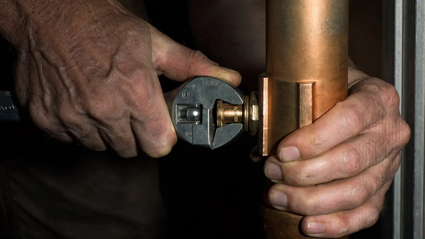 Close-up of two weathered hands in a dark cramped space, one steadying a copper pipe while the other tightens a fitting with a wrench
